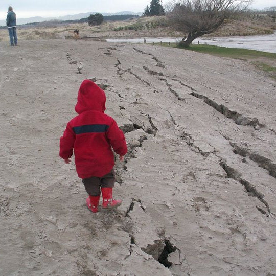 Hummingly supporting kids after floods. Child in a red coat walking on a cracked mud surface with a person in the distance.